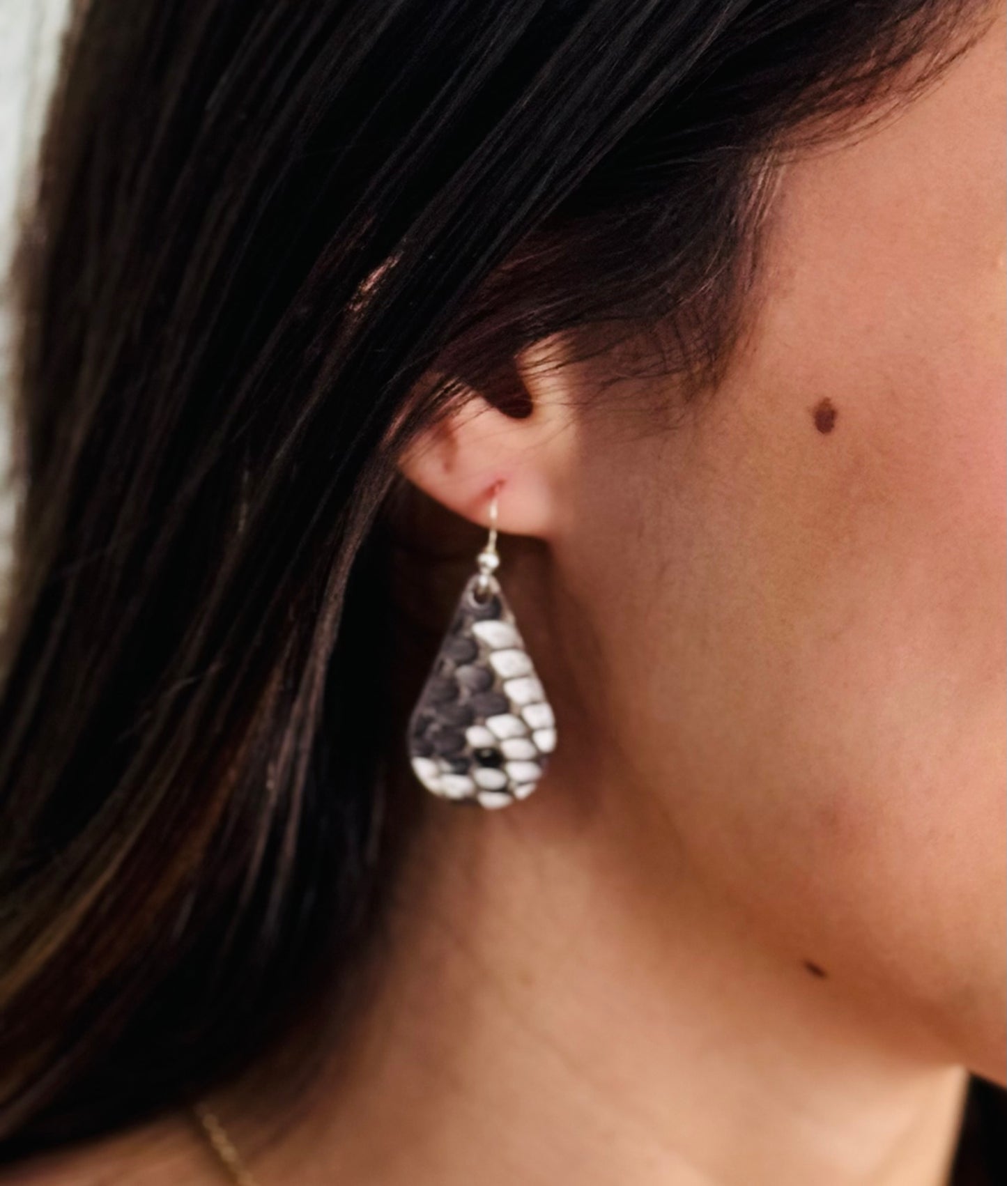 A close-up image of a woman's ear wearing a teardrop-shaped earring made from snakeskin.